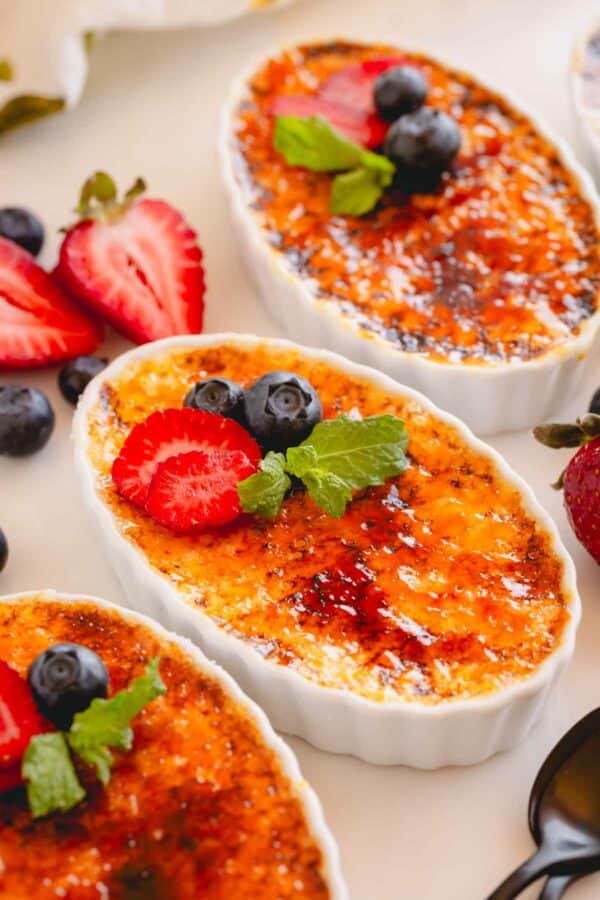 Four ramekins of classic crème brûlée topped with fresh berries and mint leaves.